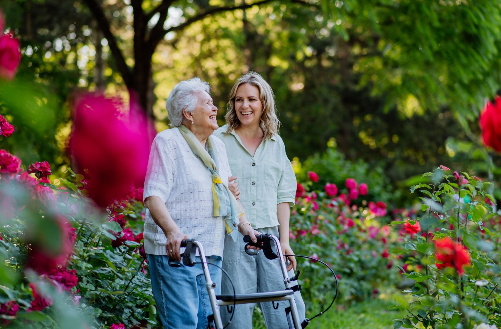 Older adult is enjoying a stroll through a garden with their adult child, both smiling, while surrounded by red flowers.