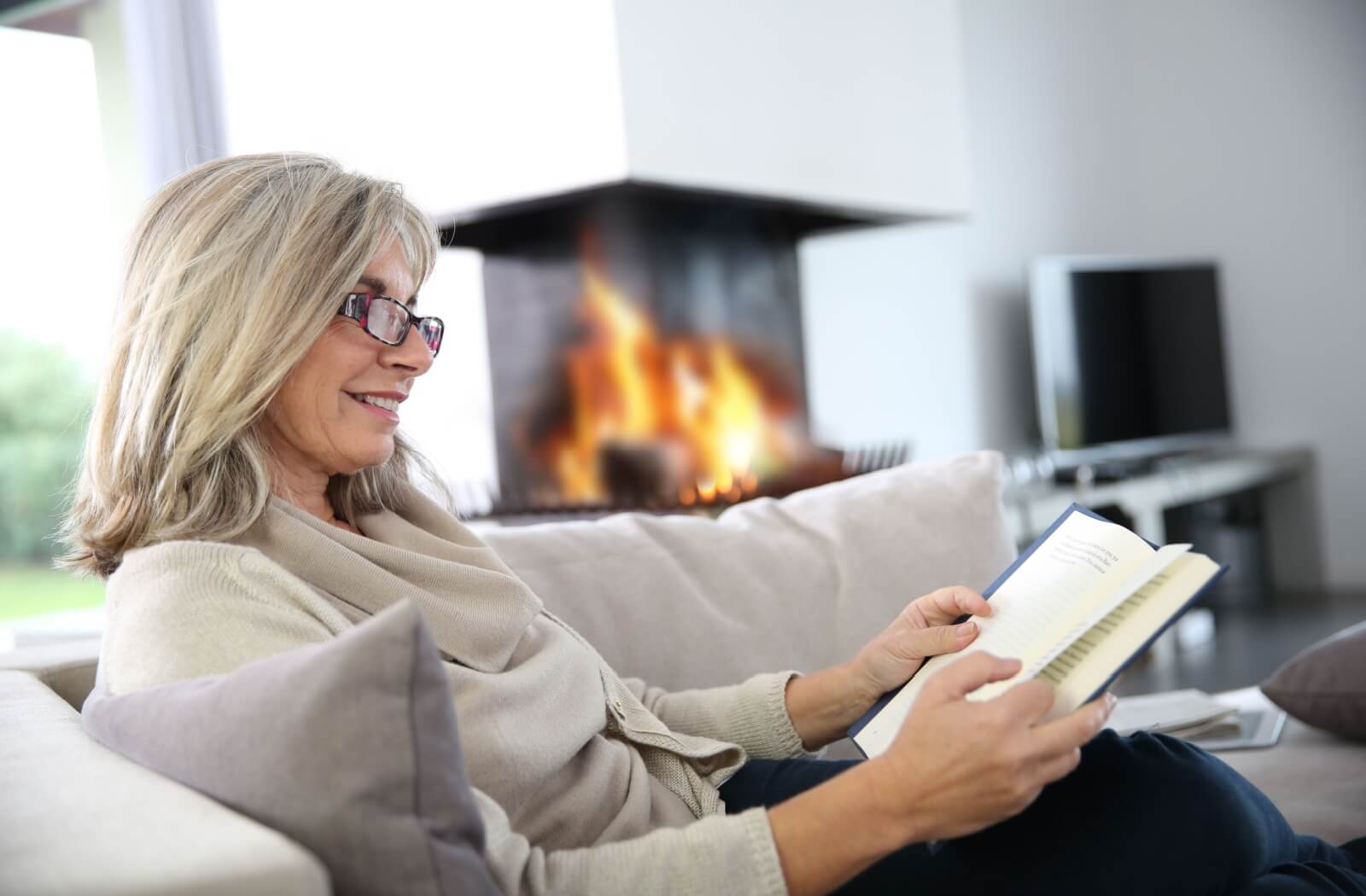 A senior enjoys a book while relaxing on their couch in front of a lit fireplace