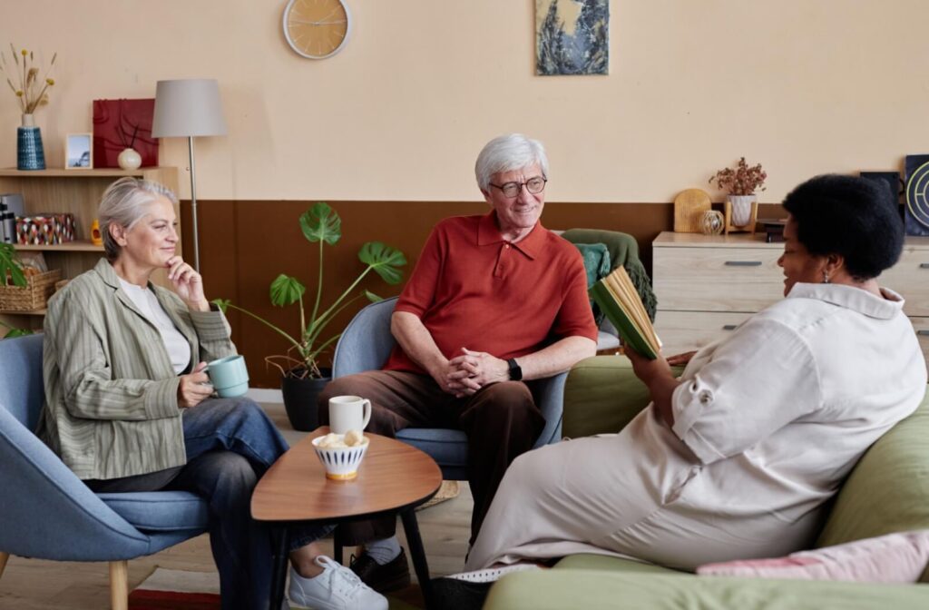 Two seniors in a communal area in memory care listen to their friend read a favorite passage from a book out loud