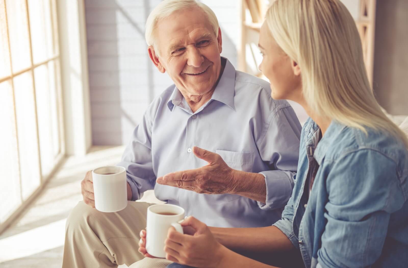 An adult child and their parent with dementia drinking coffee and chatting in front of a window in memory care.