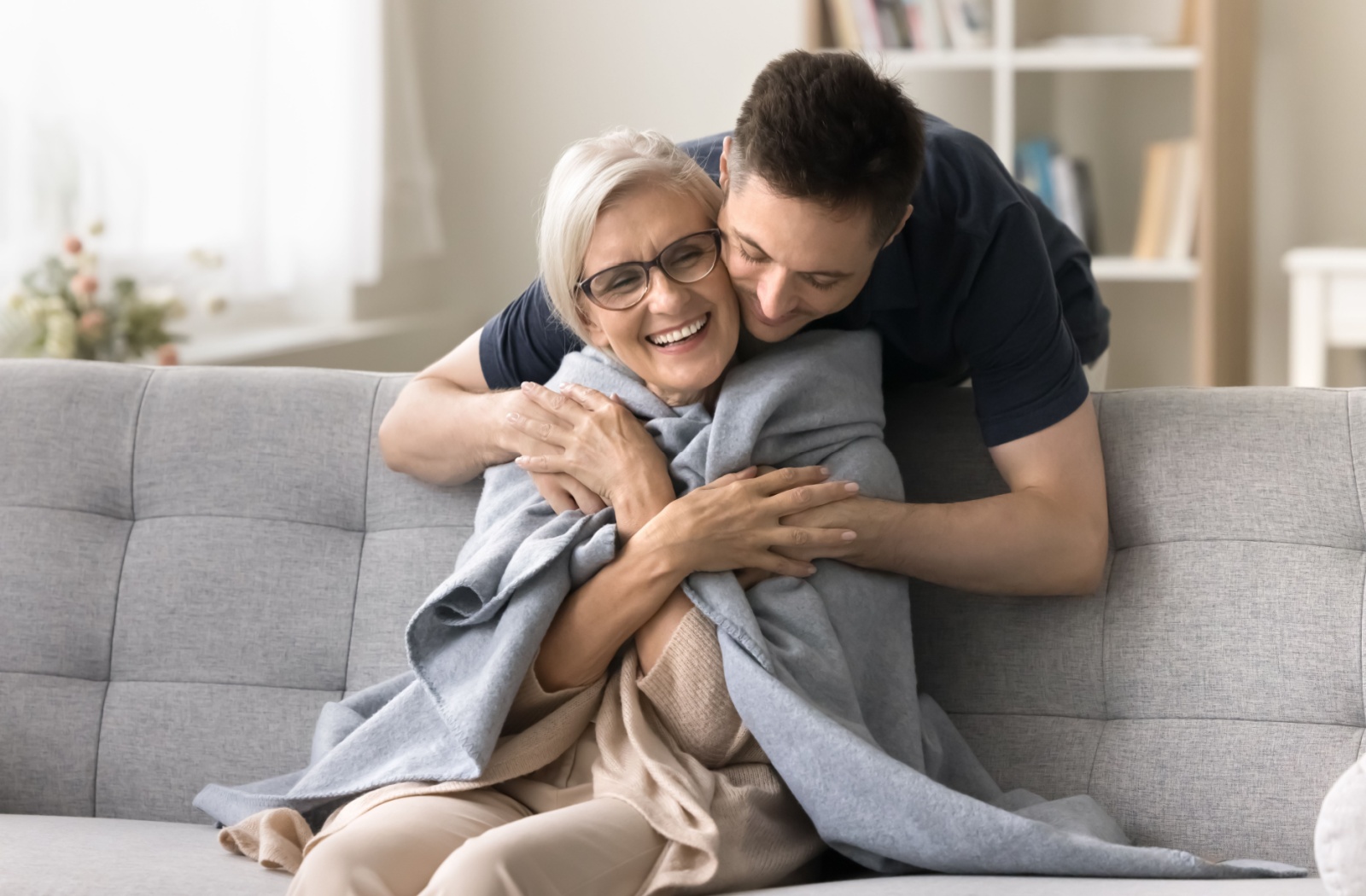 An adult hugs their parent over the shoulders after wrapping a blanket around them during a memory care visit
